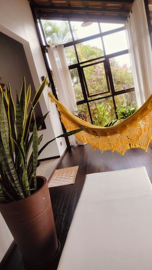 Quarto com rede, planta em vaso, janela com vista para o verde e uma mesa branca.