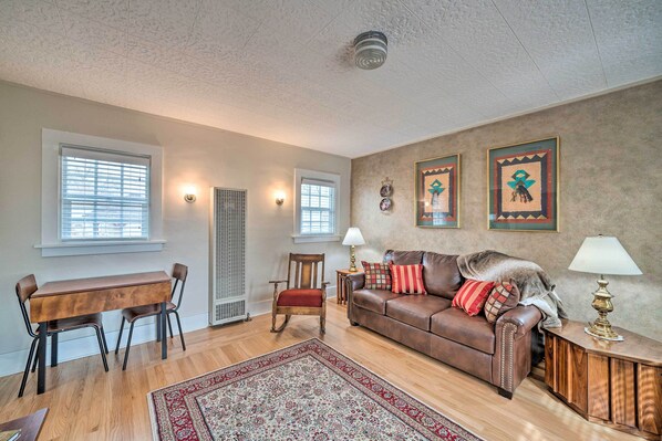 A living room with a wooden dining table, two chairs, a brown leather sofa, a red and white patterned rug, and two wall-mounted lamps. - Well-equipped Cody Cottage: 2 Mi to Cody Rodeo! (Cody)