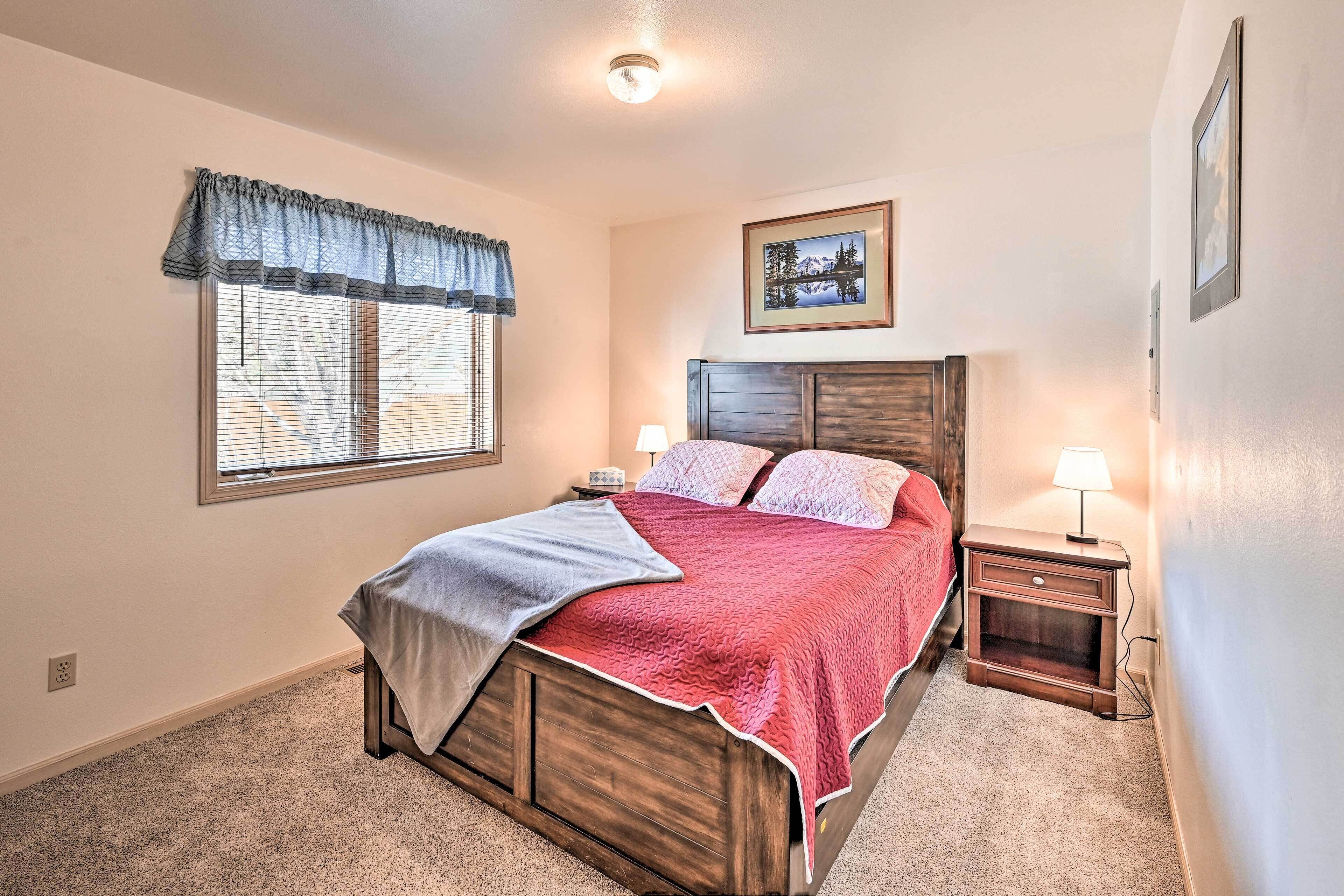 A bedroom with a wooden bed, two bedside tables with lamps, a window with blinds, and a framed picture on the wall.