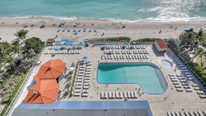 Vue aérienne d’un complexe hôtelier de bord de mer, avec une piscine, des chaises longues et des parasols.