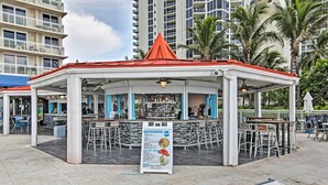 Un espace bar extérieur abrité, doté de plusieurs types d’aménagement pour s’asseoir, d’un panneau affichant le menu et offrant une vue sur les bâtiments environnants.