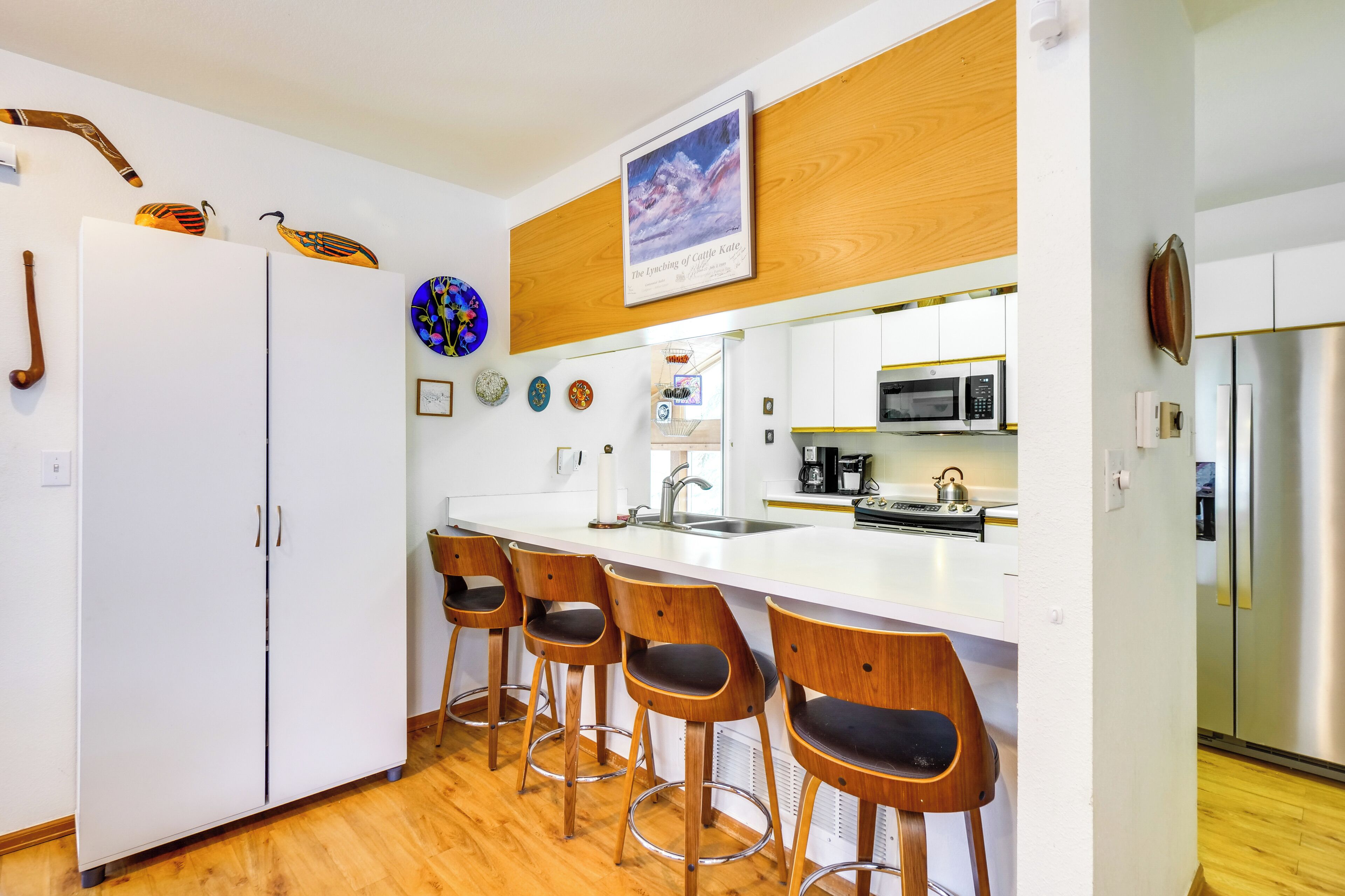 A modern kitchen with white cabinets, a stainless steel refrigerator, a microwave, and a coffee maker. There are four wooden bar stools with black cushions.