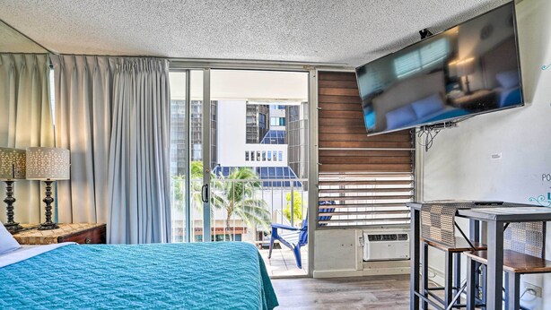 A hotel room with a bed, a TV mounted on the wall, a balcony with a chair, and a table. - Centrally Located Vacation Rental in Waikiki! (Honolulu)