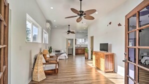 A spacious living area with hardwood flooring, a dining table, chairs, a sofa, a TV, and a wooden cabinet. There are two ceiling fans and a large window providing natural light.