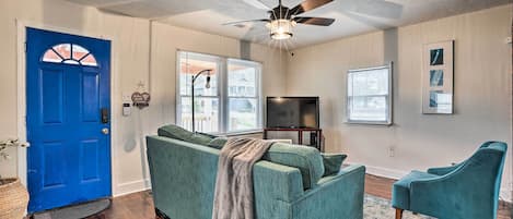 A living room with a blue door, a ceiling fan, a green sofa, a television, and a window with blinds.
