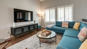 A living room with a dark wood floor, a blue sofa, a glass coffee table, a black entertainment unit, and a large TV. - Modern North Potomac Home w/ Furnished Deck! (Gaithersburg)