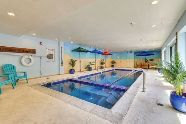 An indoor swimming pool with clear blue water, surrounded by a tiled deck. There are lounge chairs and umbrellas, and the walls are painted with a beach scene.