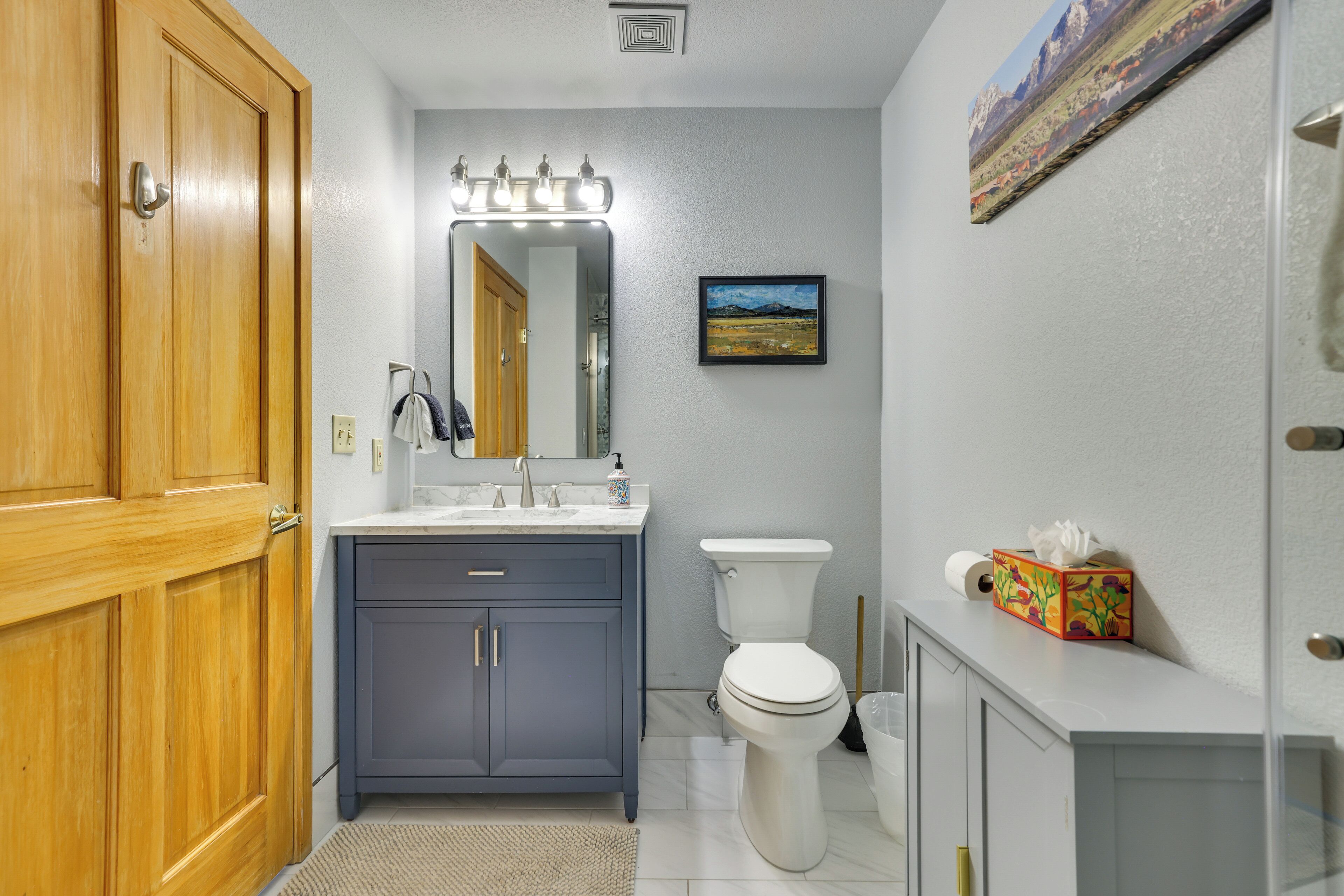 A bathroom with a wooden door, a sink with a mirror above it, a toilet, and a framed picture on the wall.