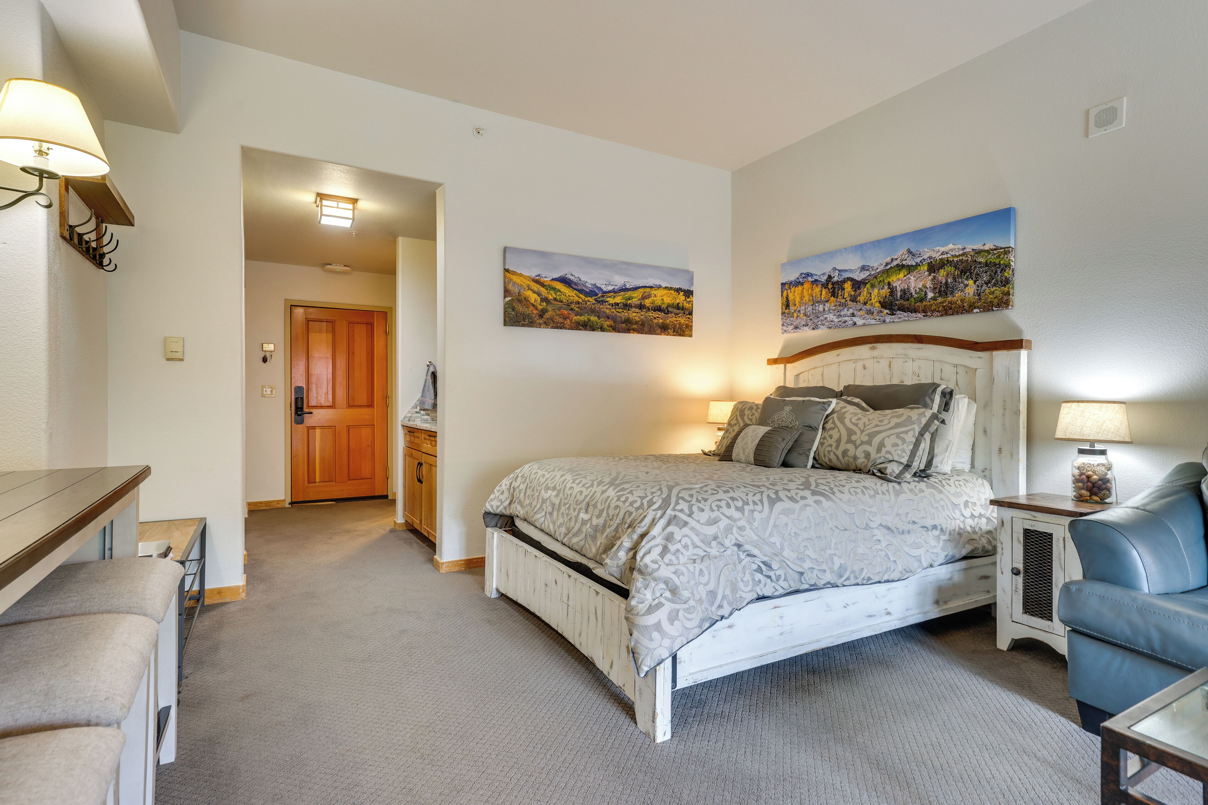 A bedroom with a bed, a desk, and a chair. There are two paintings on the wall, a door, and a light fixture.