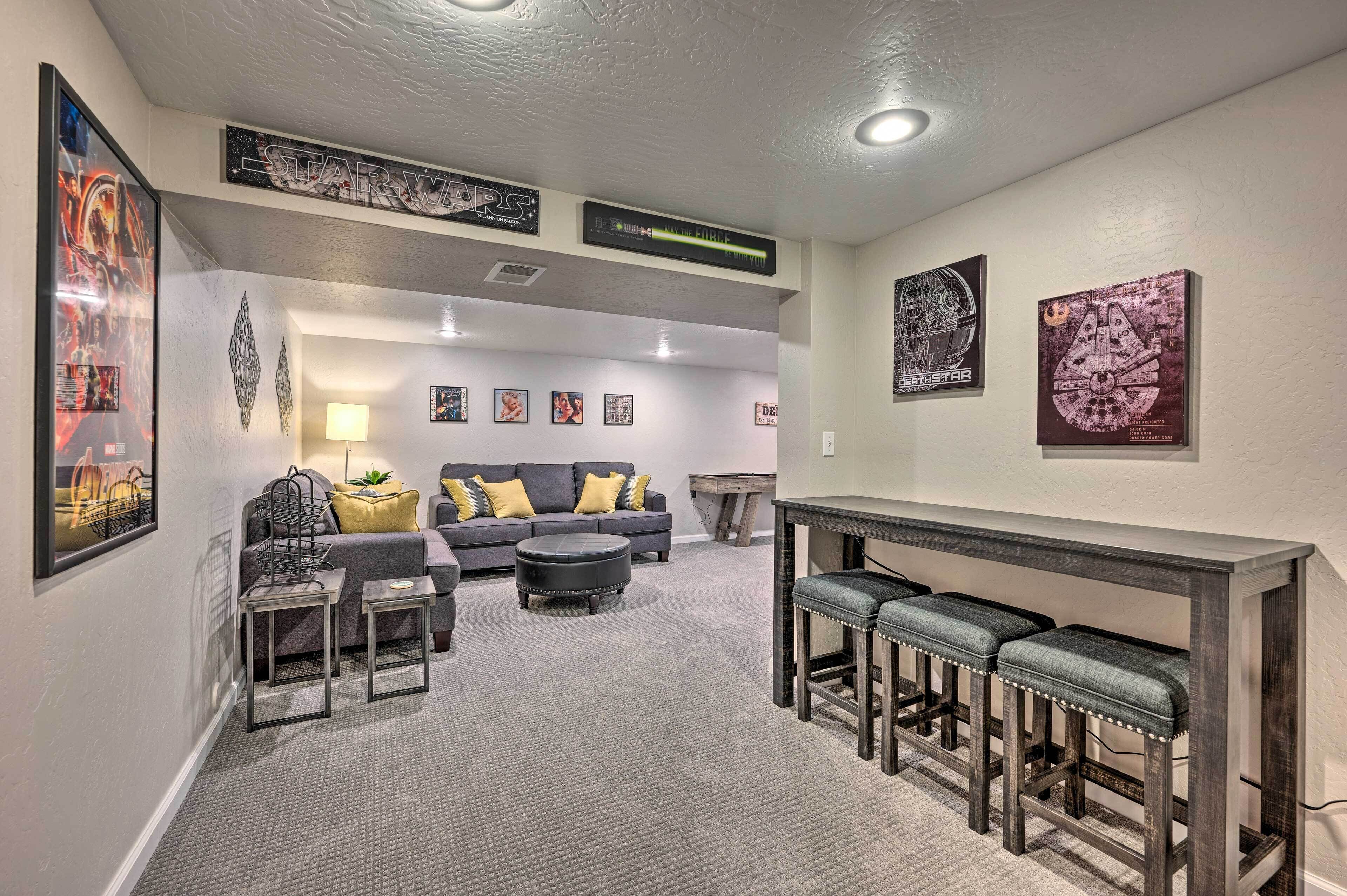 Un salon comprenant un canapé, un pouf et des tabourets de bar. Les murs sont ornés de posters encadrés et le plafond est doté d’un luminaire.
