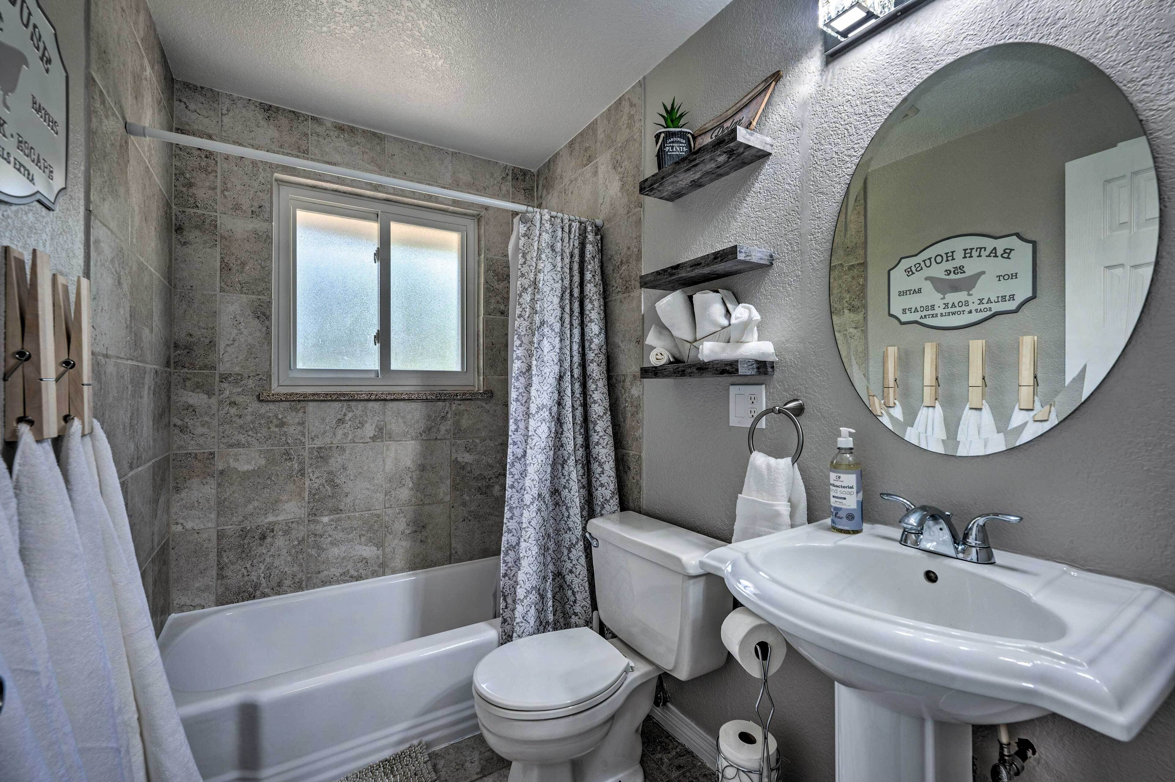 Un baño con bañera, inodoro y lavabo. Hay una ventana con puerta de vidrio esmerilado, una cortina de ducha y un espejo con un cartel sobre el lavabo.