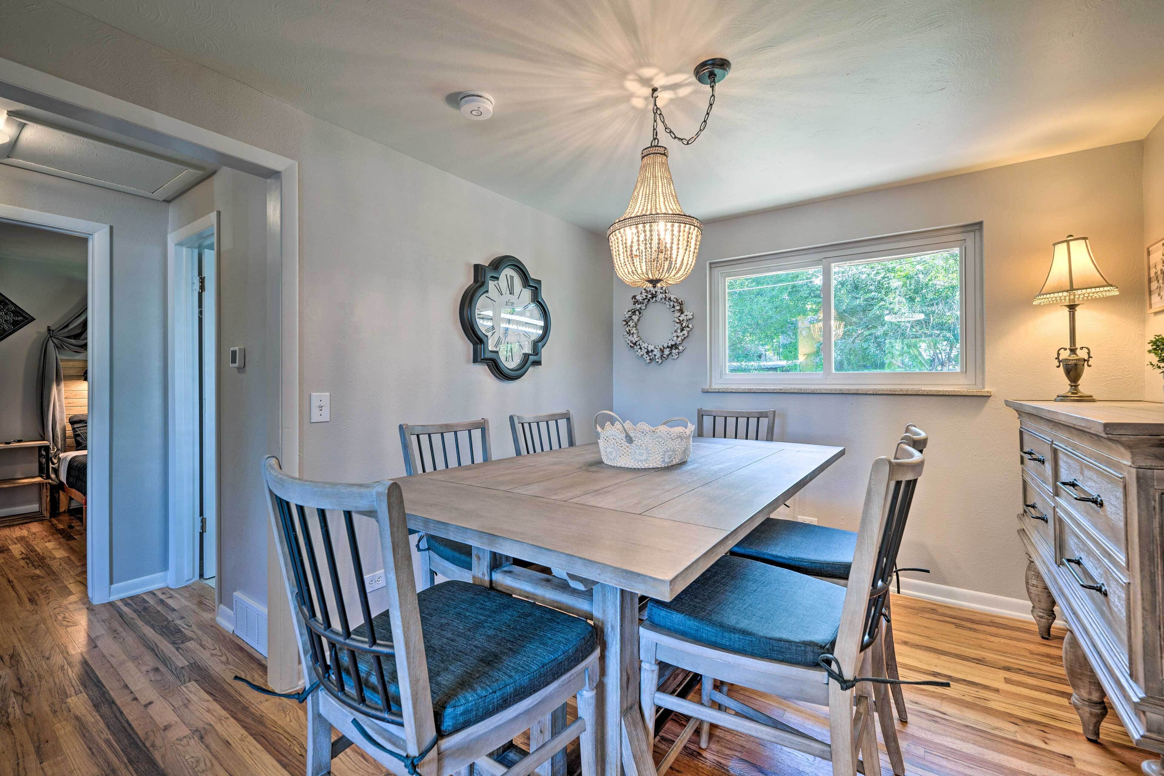 A dining area with a wooden table and chairs, a chandelier, and a window with a view of greenery.