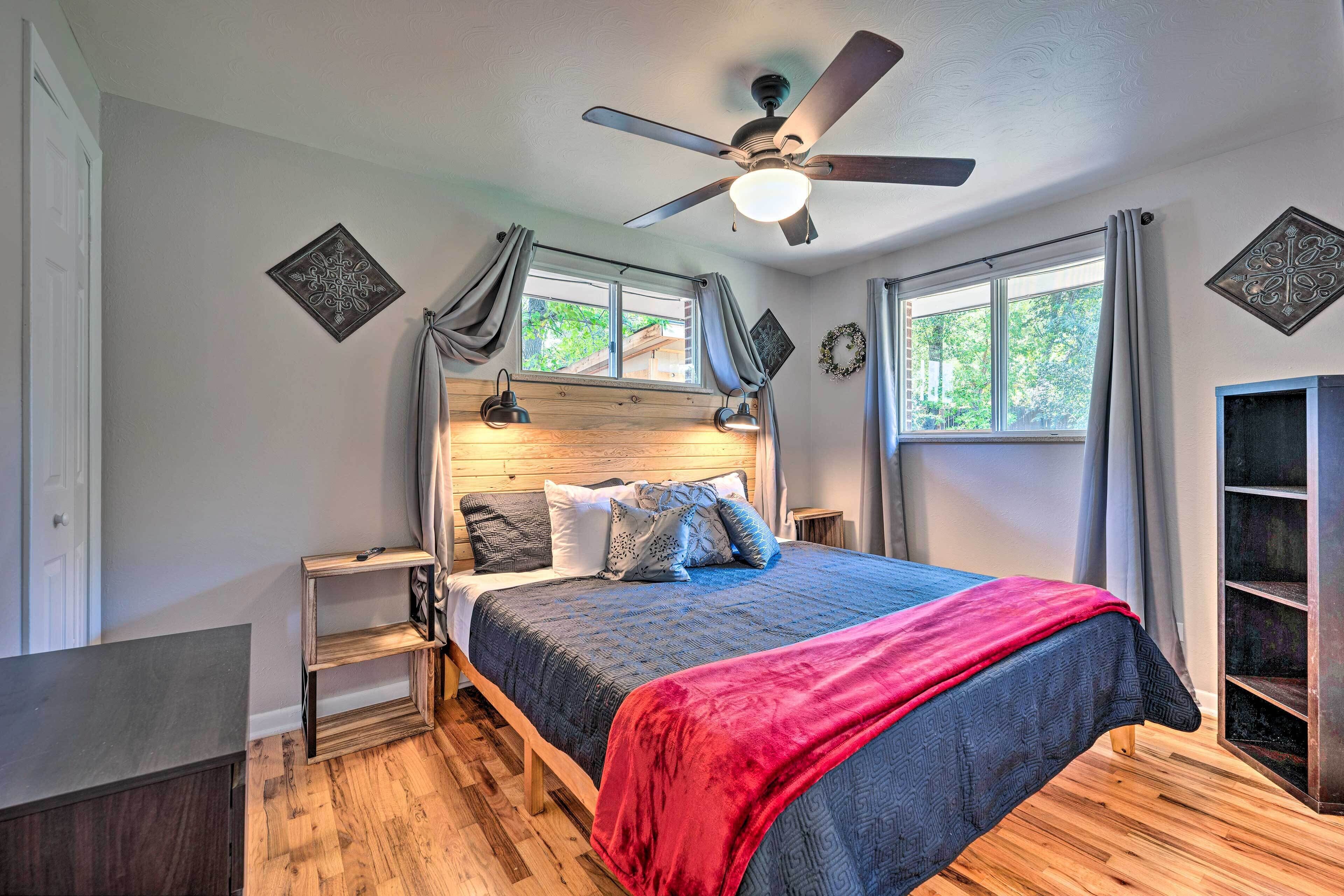 Une chambre à coucher avec un lit, un plancher en bois, un ventilateur de plafond, une fenêtre avec des rideaux et des œuvres d’art décoratives au mur.