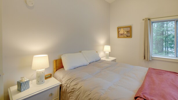 A bedroom with a bed, two bedside tables with lamps, a nightstand with a tissue box, a window with a view of trees, and a framed picture on the wall.