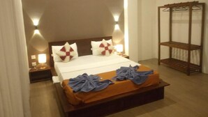 A hotel room with a bed, a nightstand, a bedside lamp, and a wooden shelf unit. - Riverdale Ridge Apartment I (Kandy)