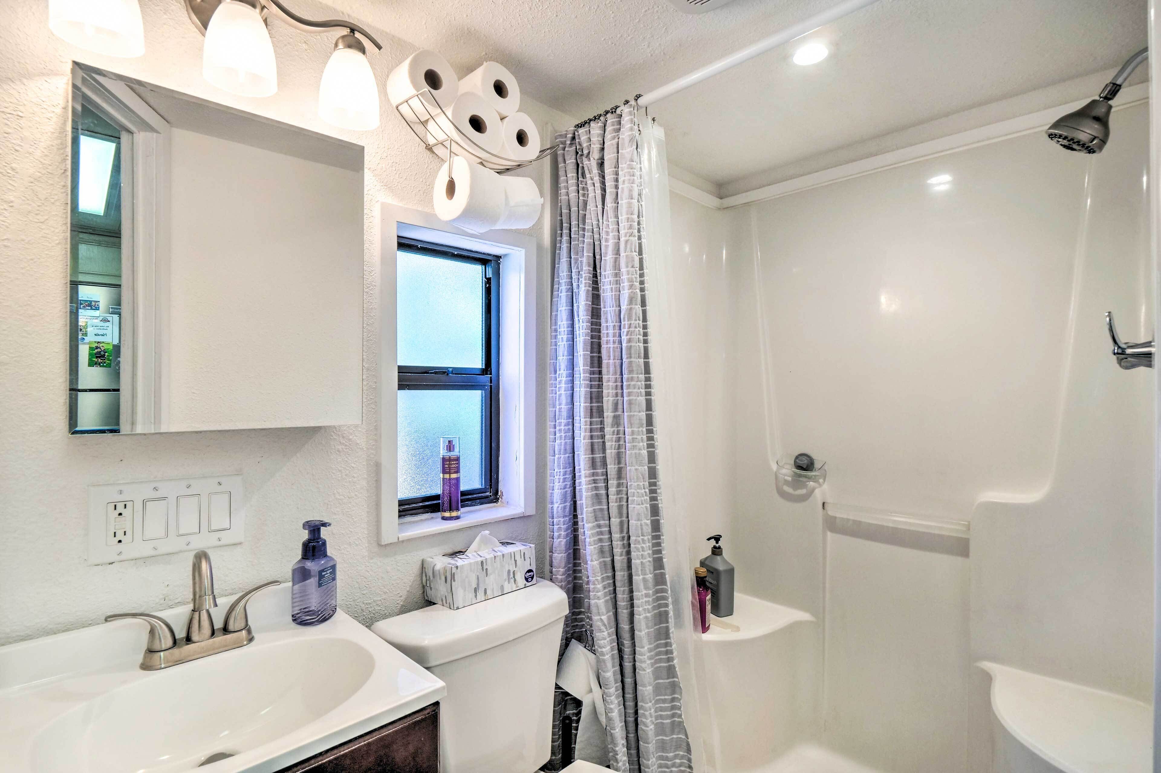 Un baño con un lavamanos blanco, un inodoro, una ducha con puerta de cristal y un ventanal con cortina.