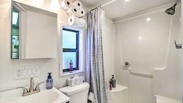 Un baño con un lavamanos blanco, un inodoro, una ducha con puerta de cristal y un ventanal con cortina.