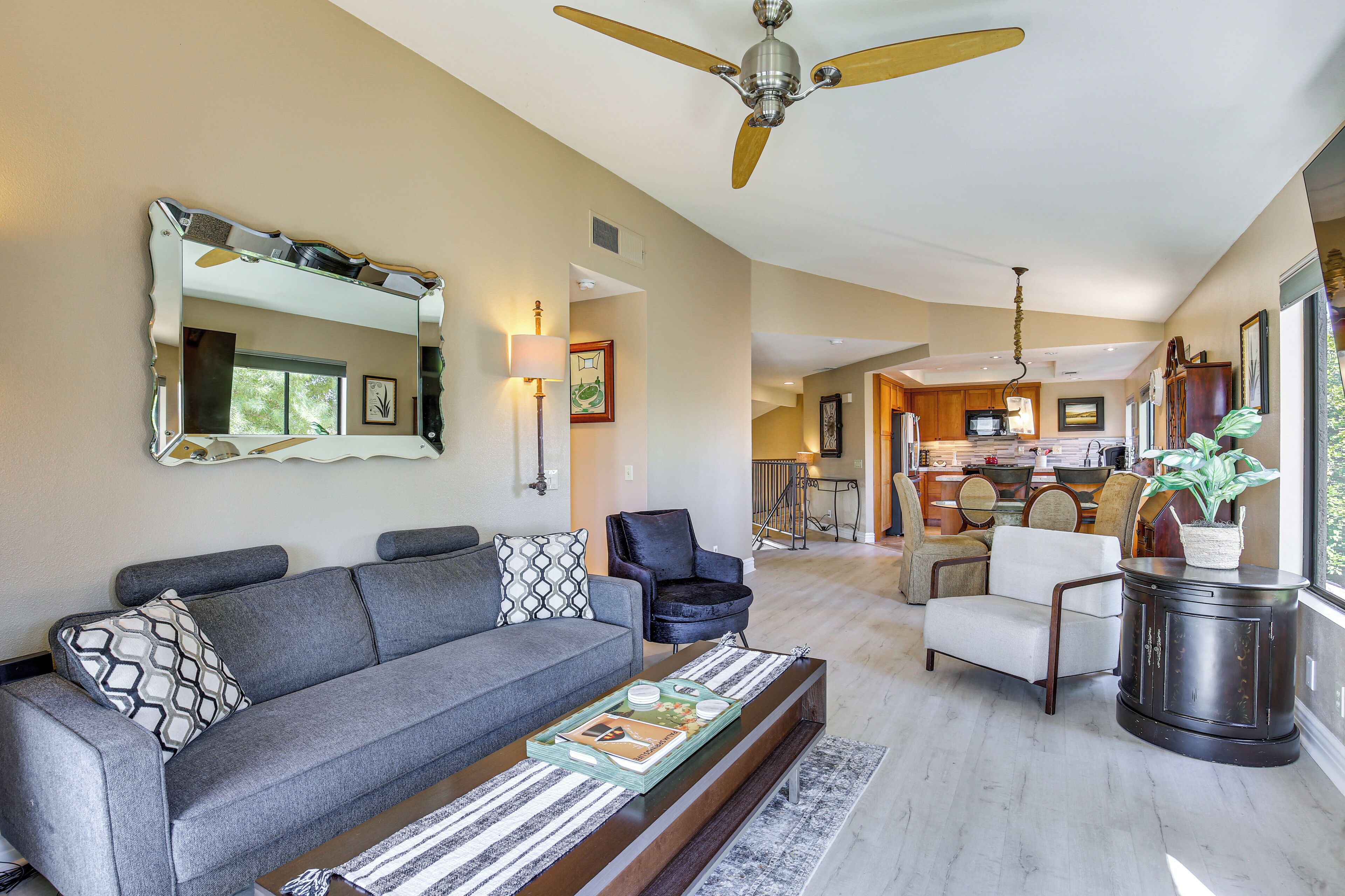A spacious living area with a grey sofa, a coffee table, and a large mirror. There is a ceiling fan and a view into the kitchen area.