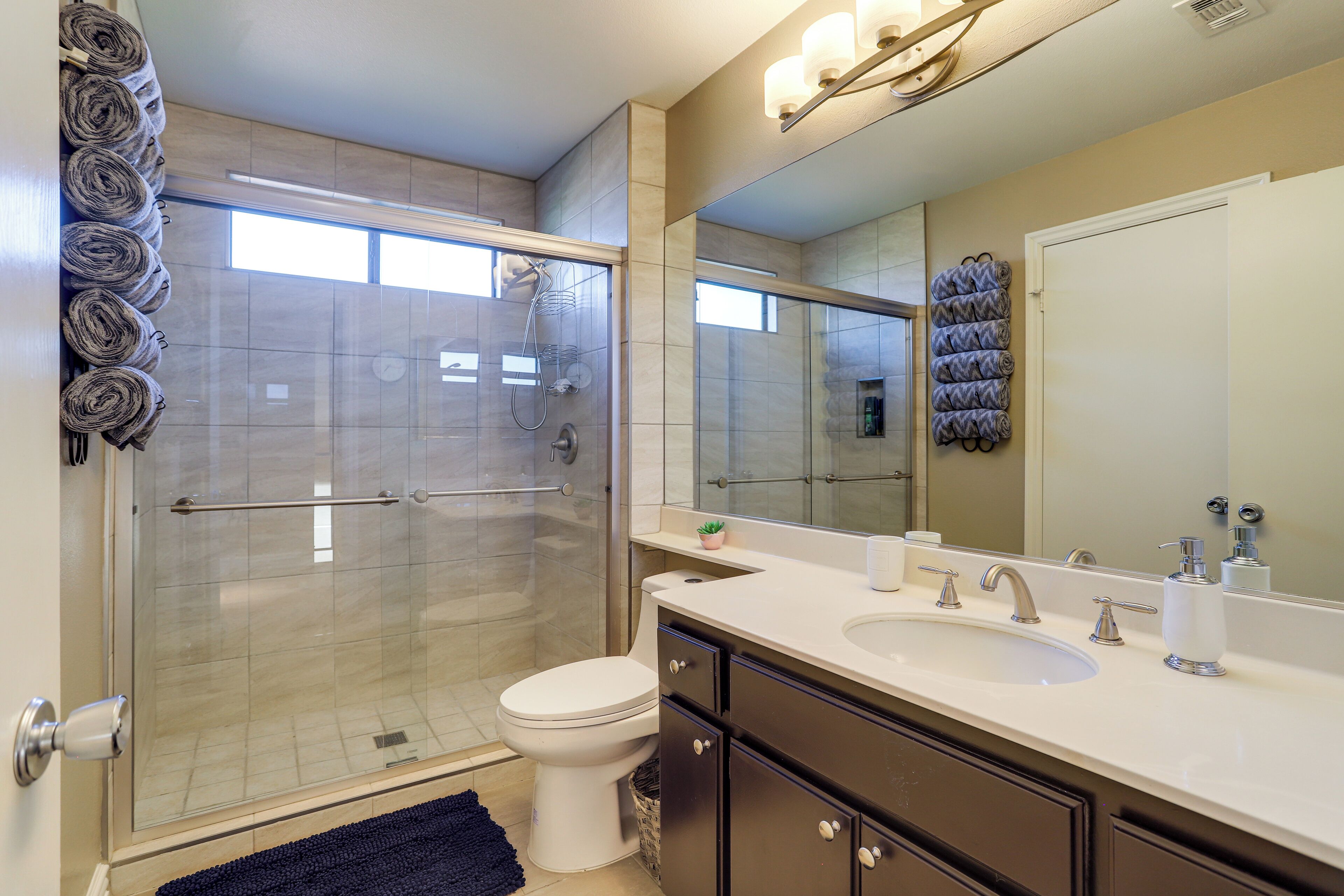 A modern bathroom with a glass shower enclosure, a white toilet, a sink with a mirror above it, and towels neatly rolled on a rack.