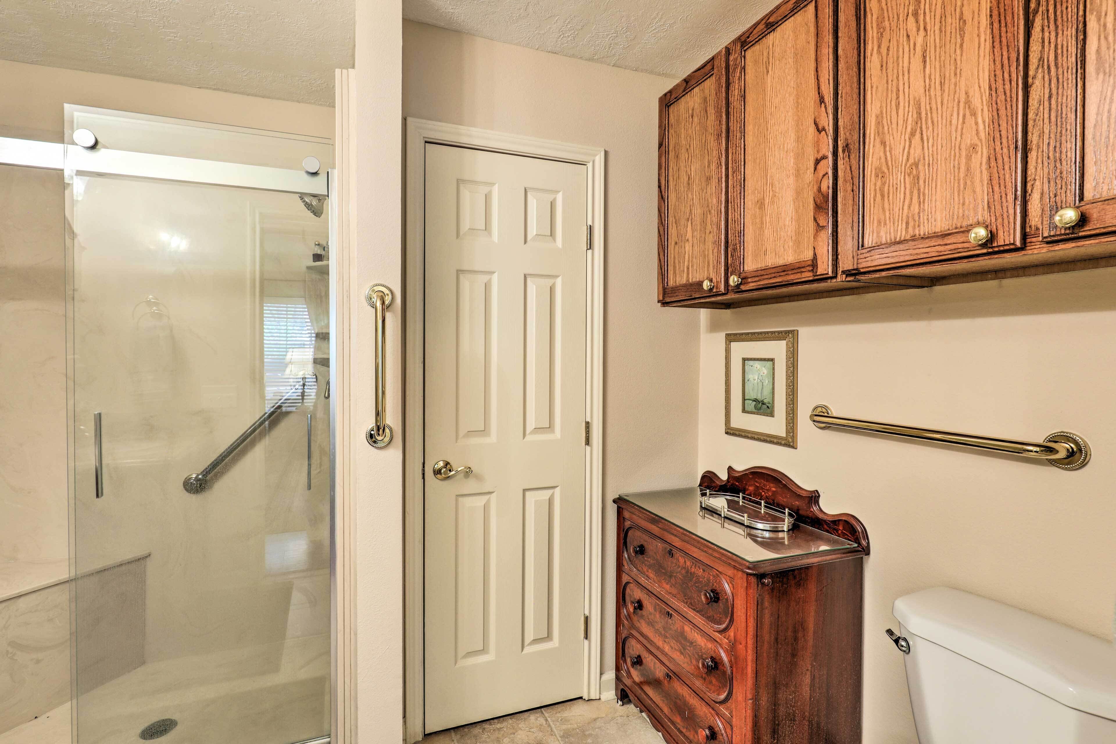 A bathroom with a glass shower enclosure, a white door, wooden cabinetry, a small wooden dresser, and a white toilet.