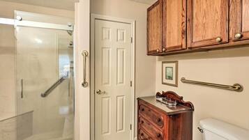 A bathroom with a glass shower enclosure, a white door, wooden cabinetry, a small wooden dresser, and a white toilet.