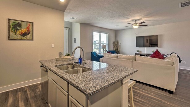 A modern kitchen with granite countertops, a double sink, and a dining area with a white sofa and a TV. - College Station Townhome w/ Furnished Patio! (College Station)