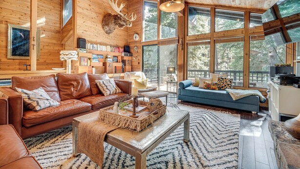 Ein gemütliches Blockhaus-Interieur mit einer großen Holzbalkendecke, einem aufgehängten Elchgeweih und Blick auf den Wald durch große Fenster.