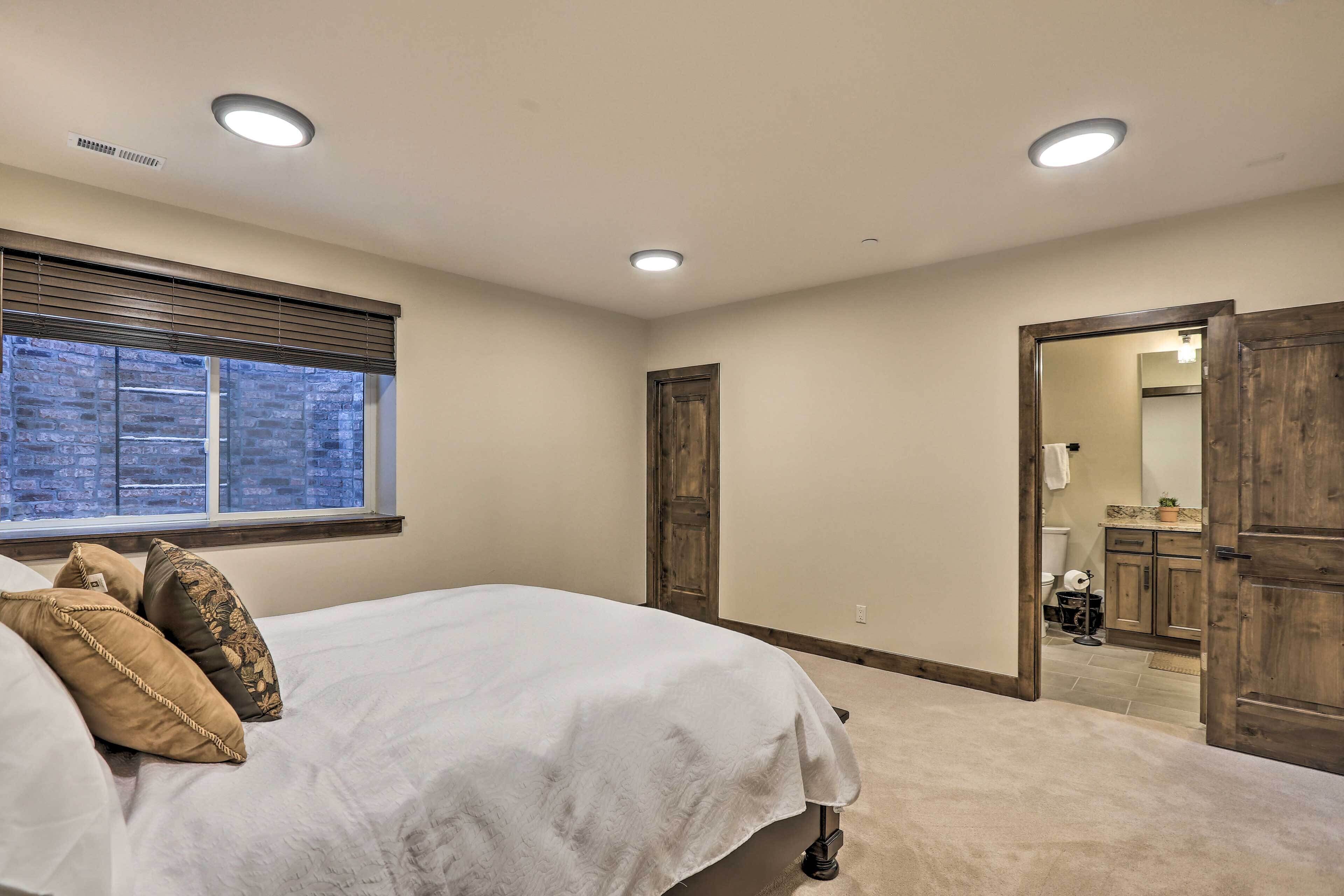A bedroom with a bed, a window with blinds, a door leading to a bathroom, and a wooden door.