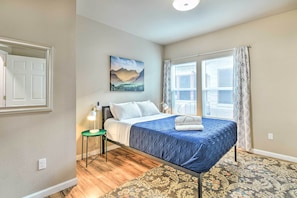 Une chambre à coucher avec un lit, une table de chevet, une lampe, une fenêtre avec des rideaux et un tableau au mur.