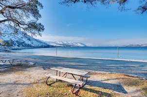 Una mesa de picnic sobre una playa arenosa, con vista despejada a un lago y montañas nevadas al fondo.
