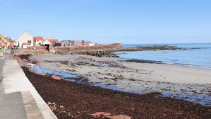 A coastal road with a beach on the right, a building with a red roof on the left, and a clear blue sky. - Experience modern coastal living at its finest in this unique one-bedroom property. Sleeps 2. (Dunbar)