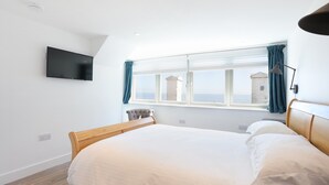 A bedroom with a bed, a TV mounted on the wall, a window with curtains, and a view of a building outside. - Experience modern coastal living at its finest in this unique one-bedroom property. Sleeps 2. (Dunbar)