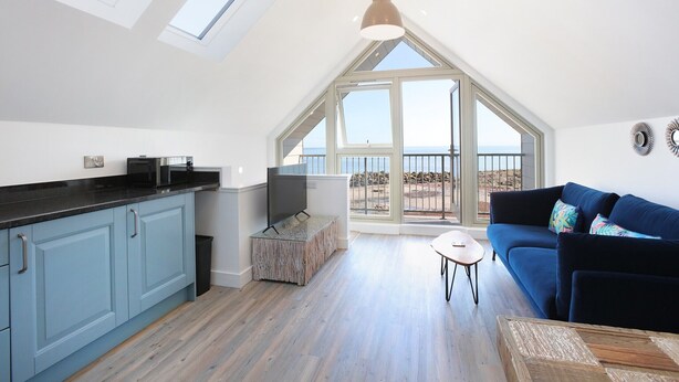 A spacious loft with a kitchen, a living area with a sofa, and large windows offering a view of the outdoors. - Experience modern coastal living at its finest in this unique one-bedroom property. Sleeps 2. (Dunbar)