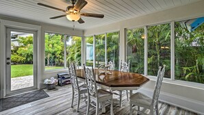 A dining area with a wooden table and white chairs, large windows with a view of a lush garden, and a ceiling fan.