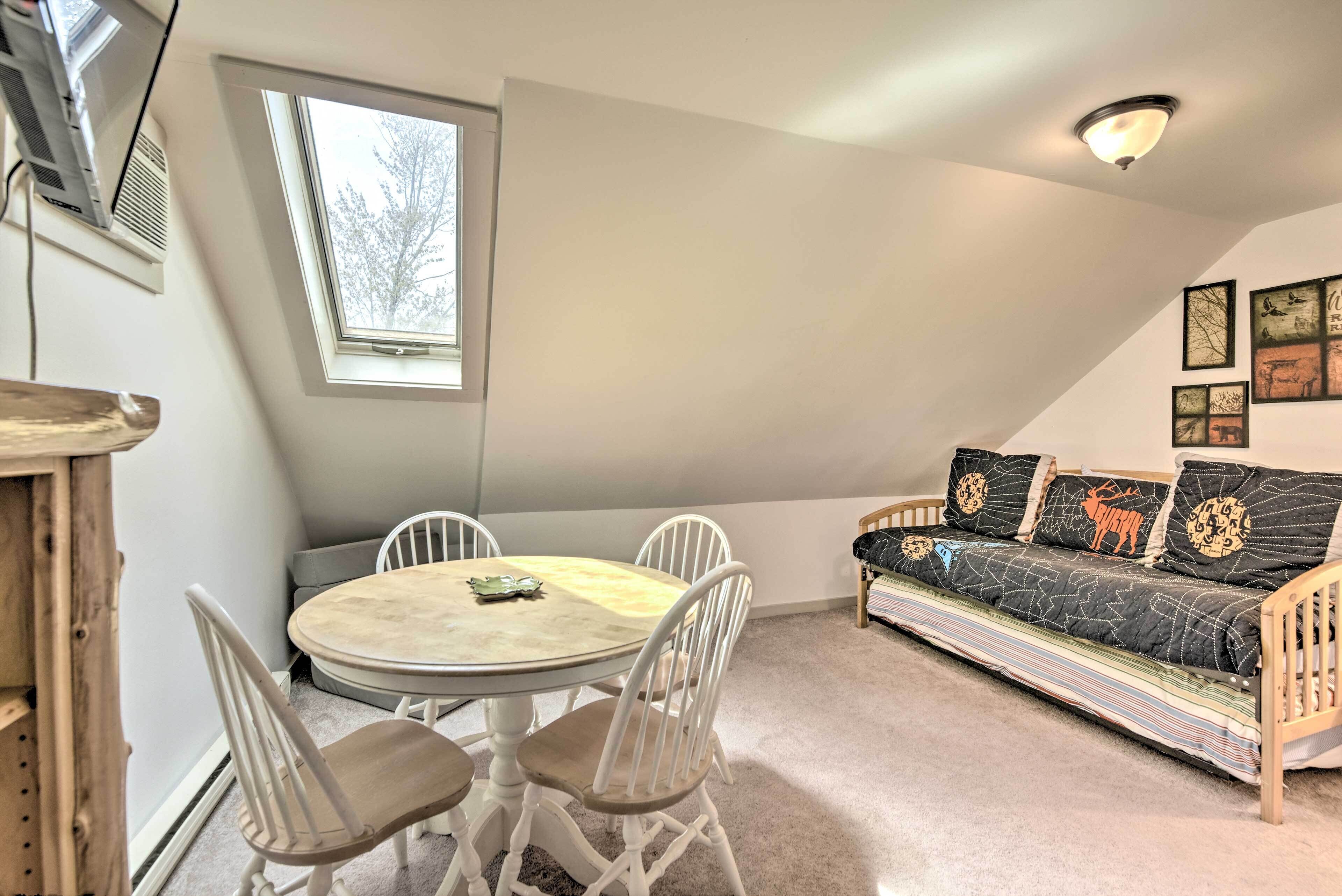 Una habitación acogedora con una mesa redonda y cuatro sillas, un sofá con cojines estampados, y un techo inclinado con tragaluz.