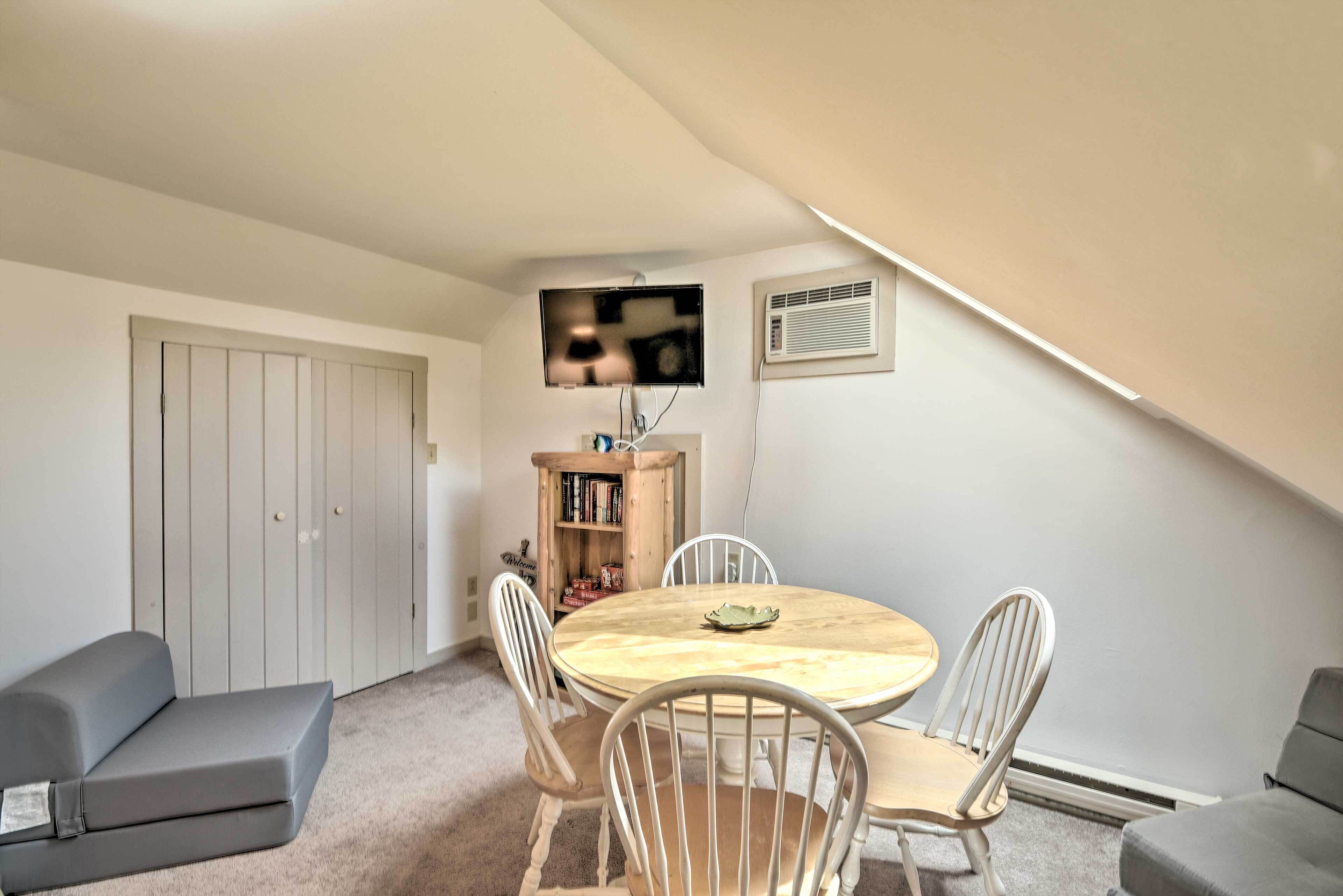 A small, cozy room with a round wooden table, white chairs, a bookshelf, and a grey sofa.