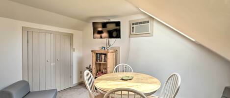 A small, cozy room with a round wooden table, white chairs, a bookshelf, and a grey sofa.