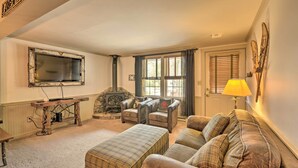 A cozy living room with a fireplace, leather armchairs, a plaid ottoman, and a television mounted on the wall. - Townhome w/ Deck: Walk to Mirror Lake & Main St! (Lake Placid)