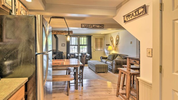 A spacious living area with a dining table, wooden chairs, a sofa, and a coffee table. A refrigerator is visible on the left, and a sign reading "LAKEFLAT" is on the wall. - Townhome w/ Deck: Walk to Mirror Lake & Main St! (Lake Placid)