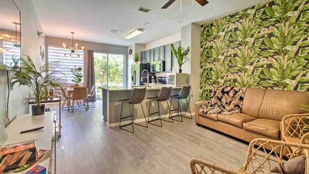 A modern kitchen with a dining area, a living room with a sofa and chairs, and a large window with blinds.