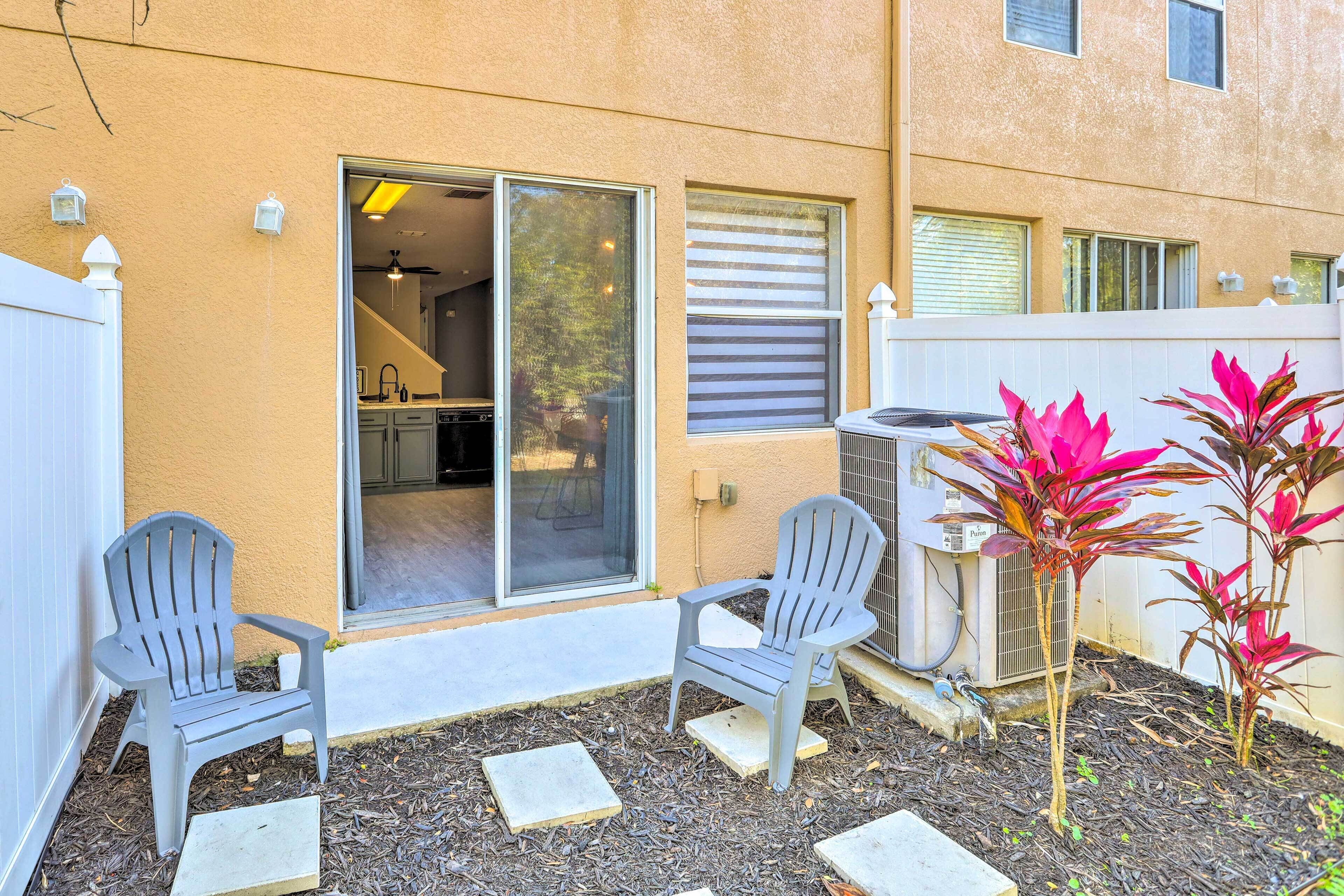 Um pátio com duas cadeiras Adirondack, um pequeno jardim com plantas de folhas vermelhas, e uma porta de correr de vidro que dá para uma cozinha.