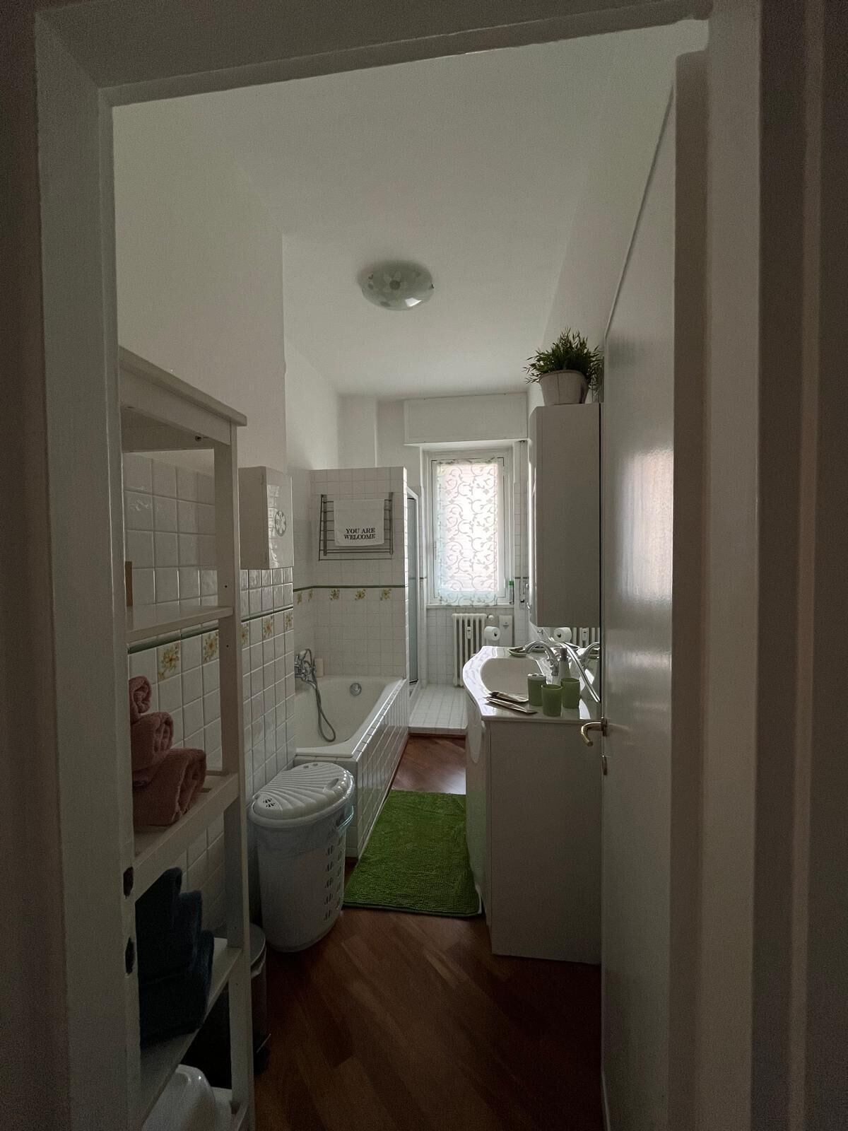 Un couloir étroit mène à une salle de bain équipée d’une baignoire, d’un lavabo et de toilettes. Un tapis vert recouvre le sol, et des serviettes sont visibles sur des étagères.