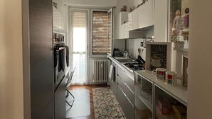 Cuina estreta amb nevera, fogó i armaris. Es veu una finestra amb persianes, i un petit moqueta al terra.
