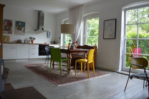 A dining area with a table and chairs, a window with curtains, and a kitchen in the background.