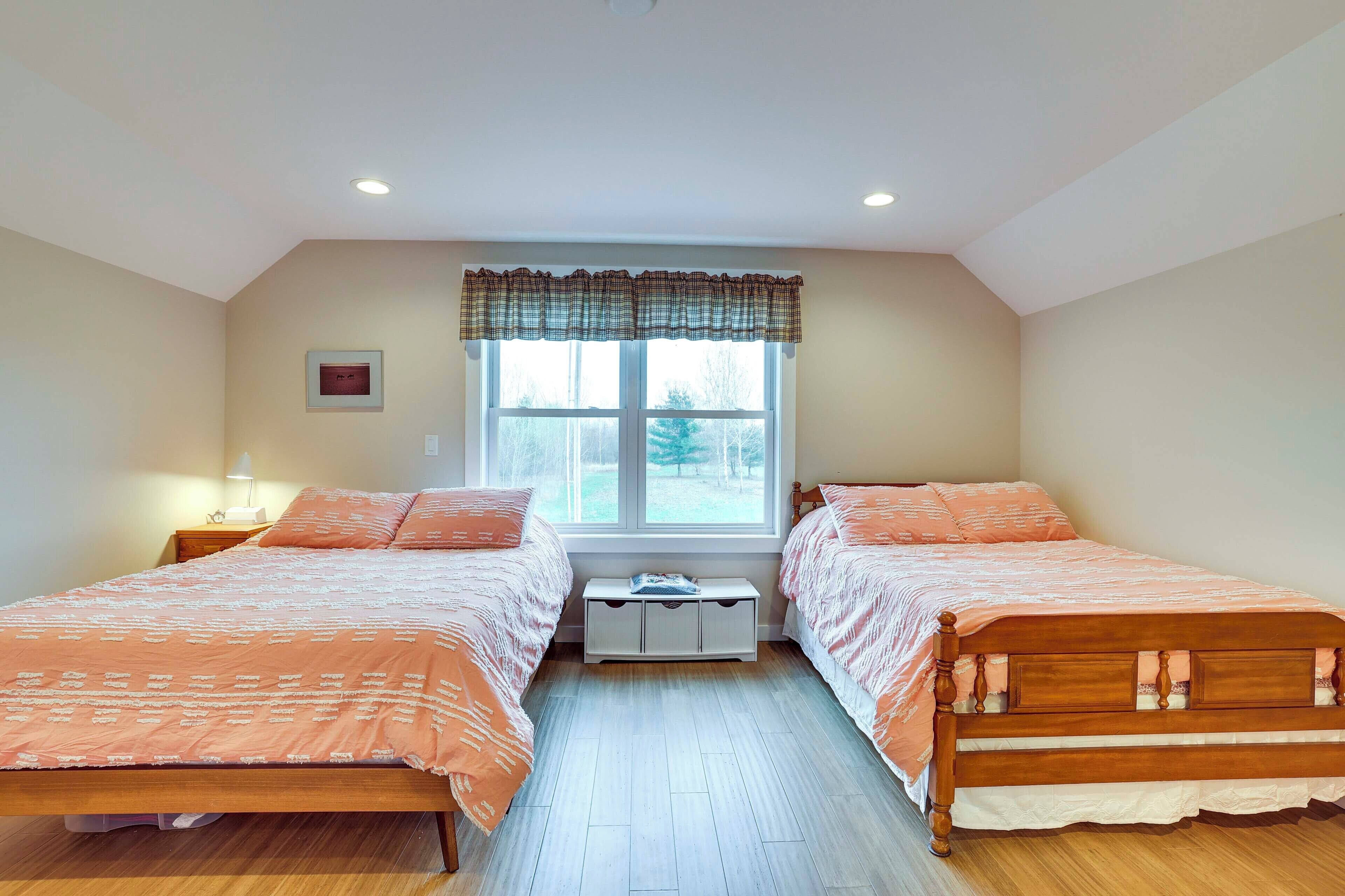 A bedroom with two wooden beds, a window with a plaid valance, a bedside table with a lamp, and a small cabinet under the window.