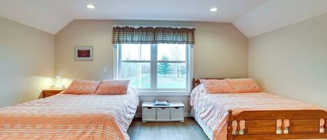 A bedroom with two wooden beds, a window with a plaid valance, a bedside table with a lamp, and a small cabinet under the window.