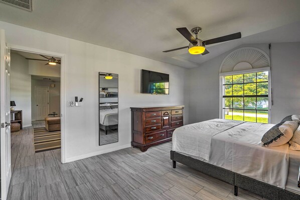 A bedroom with a bed, dresser, mirror, ceiling fan, and a window with a view of greenery.