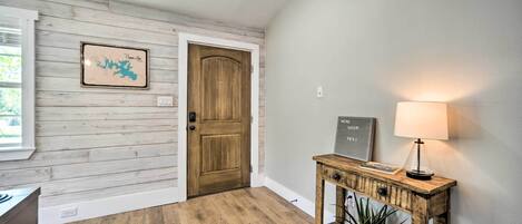 Un pasillo con una puerta de madera, un mapa enmarcado, una consola con una lámpara y una planta en maceta.