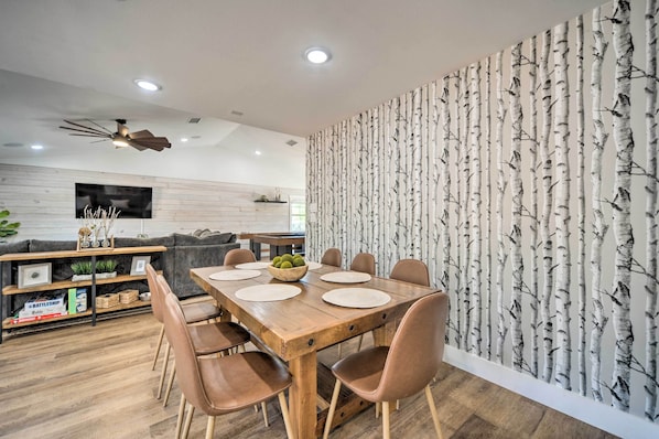 A modern dining area with a wooden table and chairs, a TV mounted on the wall, and a patterned wallpaper on the adjacent wall. - Gorgeous Cabin w/ Pergolas, Walk to River! (Canyon Lake)