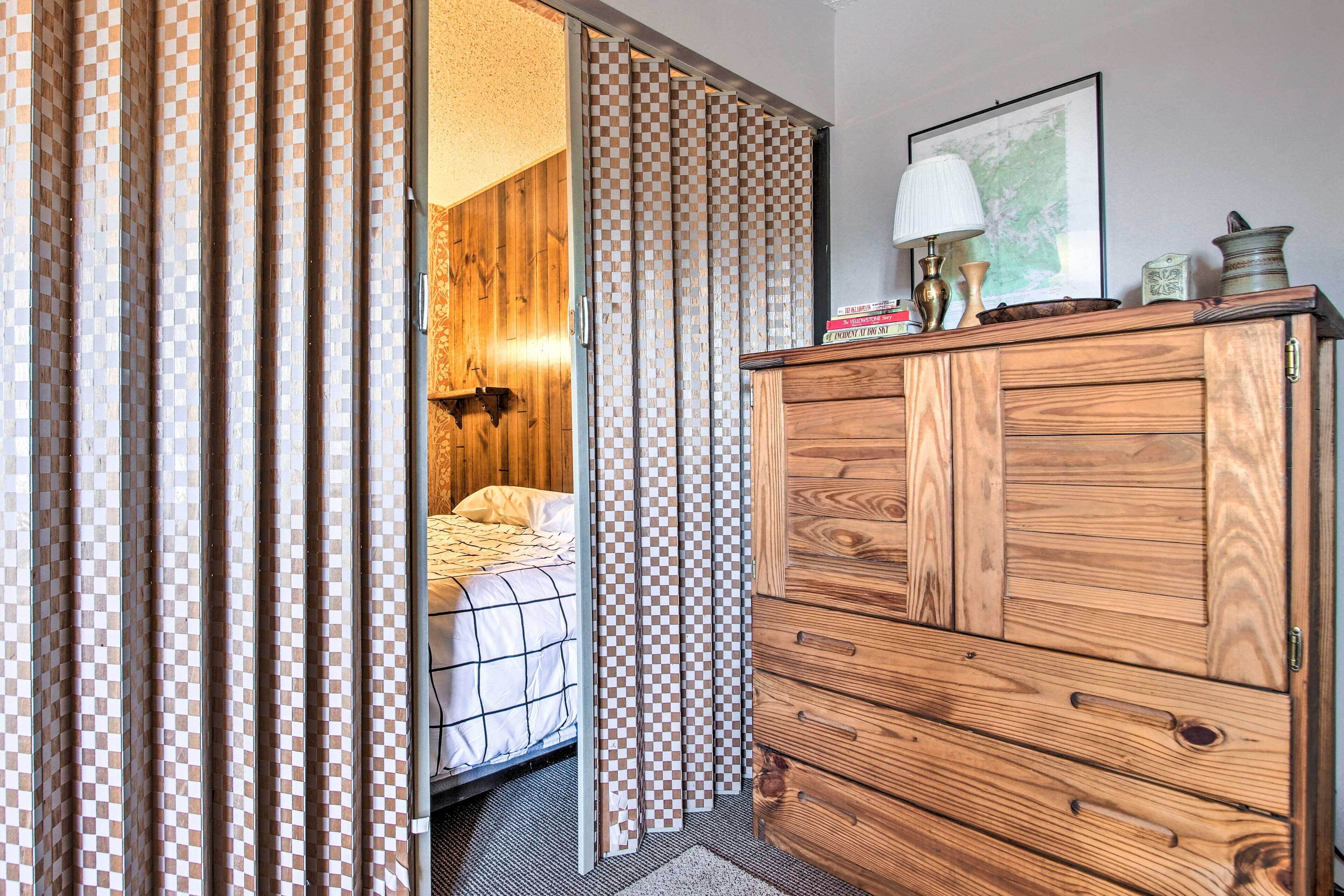 A bedroom with a wooden dresser, a bed with a checkered bedspread, and a view through a checkered curtain into another room.